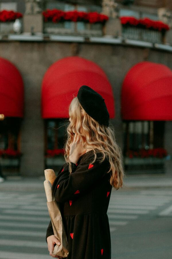 Stylish woman in black dress with red hearts holding a baguette on a street with red awnings.